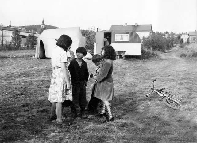 Fem barn står ihop och pratar. I bakgrunden ser man ett litet tält och en mindre husvagn. En cykel ligger bredvid barnen. Foto i svartvitt.