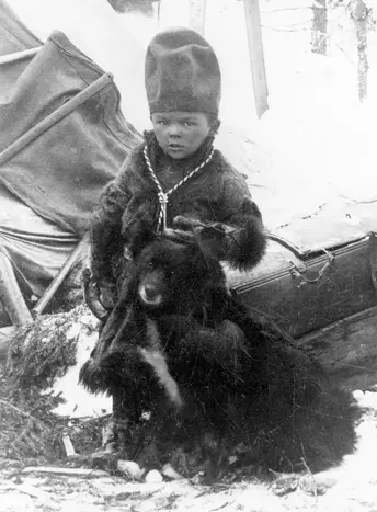 en pojke i vinterkläder står utomhus vid en sittande svart hund. Svartvitt foto.