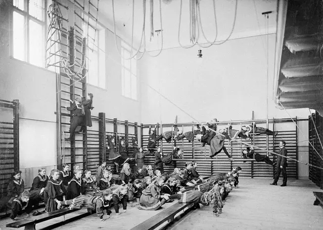 Flickor i en gymnastiksal. Några klättrar och hänger i ribbstolar och några sitter på bänkar. Foto i svartvitt.