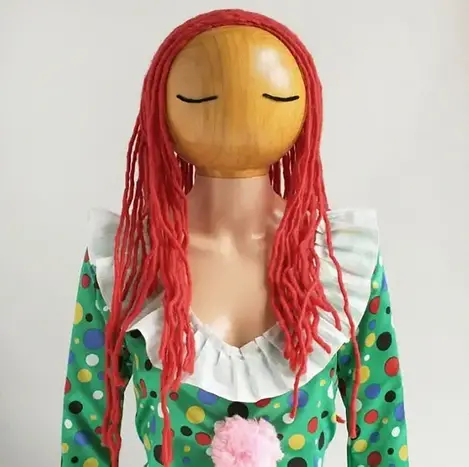 Överdel av docka i plast med trähuvud. Dockan saknar mun och näsa, har streck som ögon och rött hår av garn,. Dockan är klädd i prickig dress.