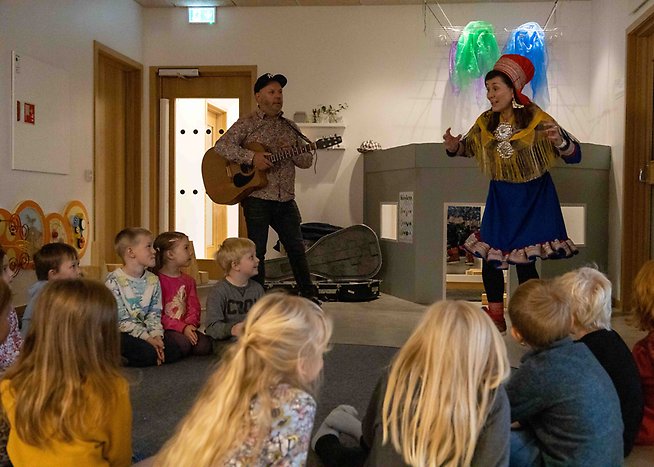 Barn sitter i ring på golvet och lyssnar till föreställning av två personer, en spelar och en sjunger i samiska kläder. 