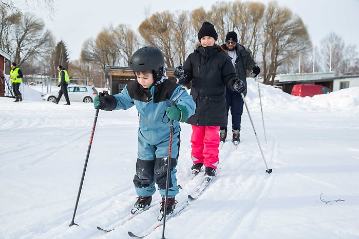 Två barn och en pappa åker skidor efter varandra. Pojken först med koncentrerad min och flickan i mitten som blickar upp mot kameran och ler och pappan kommer sist. 