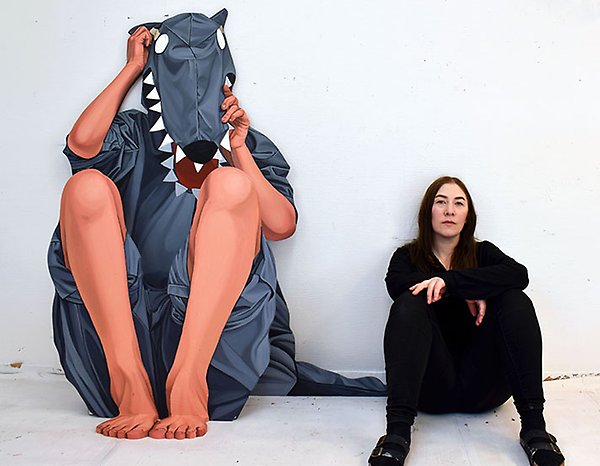  Amanda Karlsson, sitter på golvet bredvid sitt konstverk som  består av en vargliknande träfigur. 